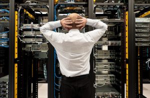 A guy with his hand on the back of his head, looking stressed at a server rack, not knowing what to do.